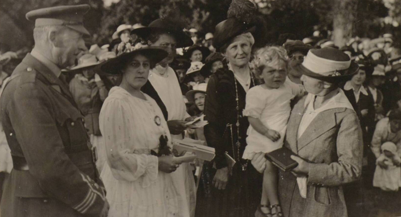 A new local organisation formed in November 1915 with the aim of supporting servicemen from Jersey being held as prisoners of war by the enemy. The Prisoner of War Bureau managed by Mrs Greg Haines opened in 3 Brook Street, St Helier to receive, process and despatch ‘comforts’ donated by Islanders for the prisoners. In July 1916, the Lieutenant Governor agreed to allow a fete at Government House on 6 July 1916 in support of the cause. This image showing performers or members of the audience at the fête possibly meeting the Lieutenant Governor’s wife.
