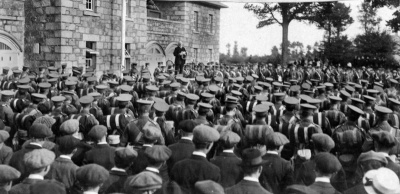 Assembled Militiamen and some civilians (possibly off-duty Militiamen) gathered at Grouville Arsenal to hear an appeal to volunteer for overseas service made by the Viscount of Jersey in October 1915. The event was part of an Island-wide campaign organised in support of Britain’s Derby Scheme - launched in the autumn of 1915 with the aim of encouraging a voluntary commitment to join-up as a last step before introducing compulsory military service.