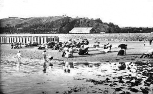 Westmount viewed from the sea. Bathing huts and people sitting in deck chairs on West Park beach. The J C A West Park Pavilion (previously known as the Tin Hut) is in the background and the walled estate of Westmount House is on top of hill